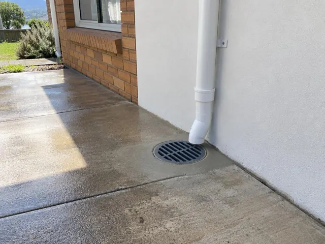 Outdoor drain grate beside a downpipe on wet concrete paving and brick wall in Wangi Wangi